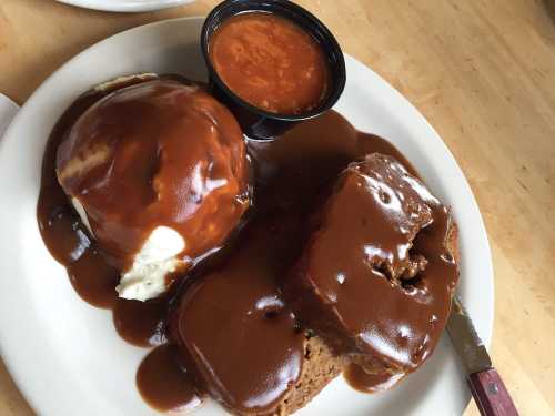 A plate with two slices of meatloaf covered in brown gravy, mashed potatoes, and a small cup of sauce.