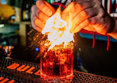 A bartender ignites a cocktail in a glass, creating a dramatic flame above the drink.