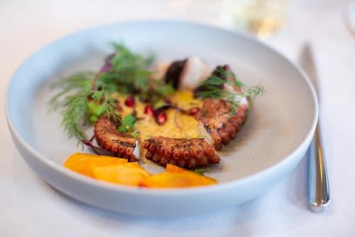 A plate of grilled octopus garnished with herbs, pomegranate seeds, and accompanied by slices of yellow fruit.