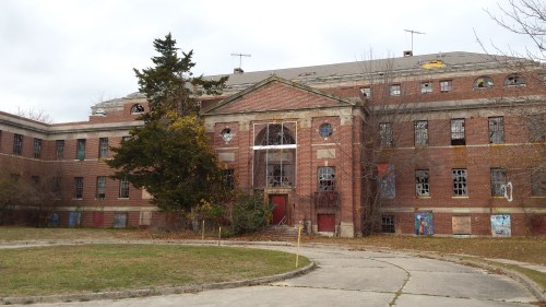 Abandoned brick building with broken windows, overgrown grass, and a large tree in front under a cloudy sky.