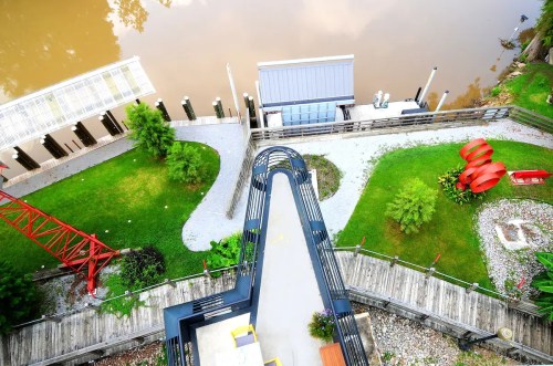 Aerial view of a riverside area featuring pathways, greenery, a red sculpture, and a dock with a building.