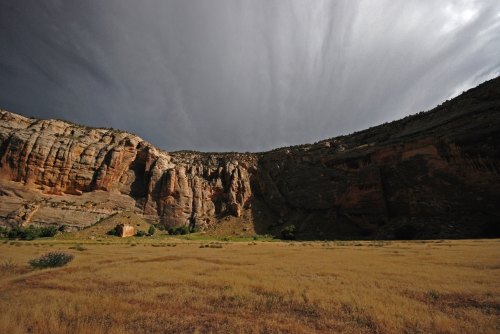 A dramatic landscape featuring towering cliffs under a cloudy sky, with a grassy plain in the foreground.