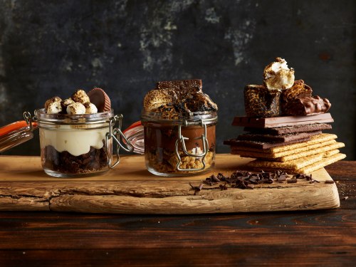 Three jars filled with layered desserts, including chocolate, graham crackers, and marshmallows, on a wooden board.