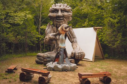 A large wooden sculpture of a smiling figure sits beside a campfire, surrounded by trees and wooden benches.