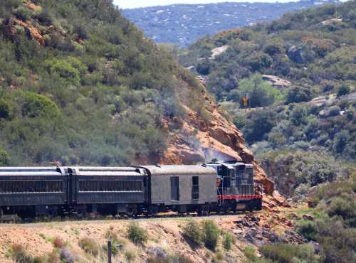 A train travels along a winding track beside a rocky hillside, surrounded by lush greenery and distant hills.