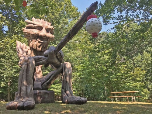 A large wooden sculpture of a troll-like figure holding a stick, surrounded by trees and a picnic table in the background.