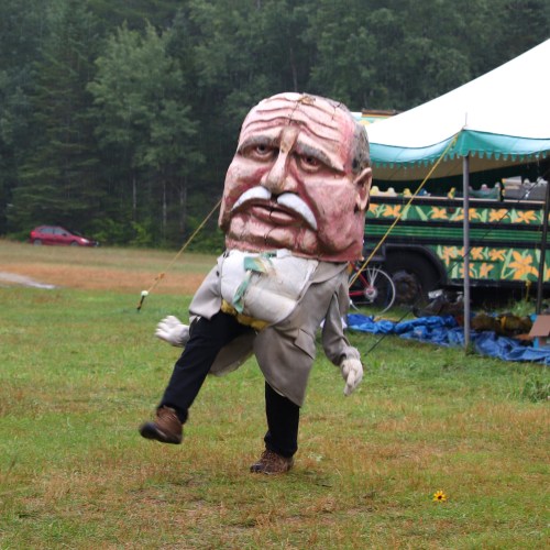 A person in a large puppet costume with a oversized head, walking on a grassy field near a tent.