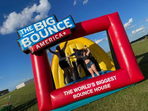 A colorful bounce house with a sign reading "The Big Bounce America" and people jumping inside against a blue sky.