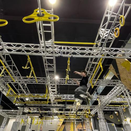 A person navigating an indoor obstacle course with yellow rings and climbing structures overhead.