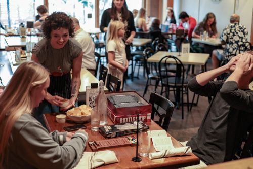 A busy restaurant scene with people dining, a waitress serving, and a group playing a game at a table.