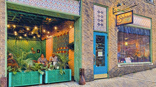 A colorful restaurant entrance with plants, featuring a sign that reads "Salam" and two people seated inside.