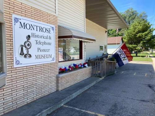 Sign for Montrose Historical & Telephone Pioneer Museum with an "OPEN" flag and decorative flowers outside.