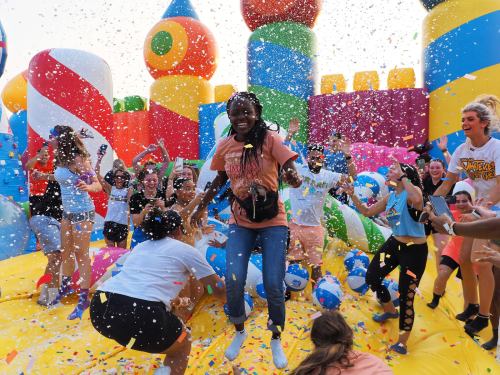 A lively scene of people jumping on a colorful inflatable structure, surrounded by confetti and beach balls.