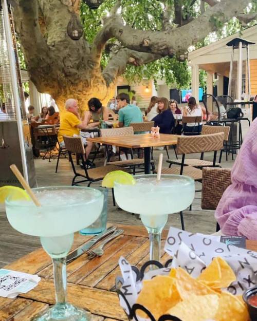 Two margaritas with lime sit on a wooden table, surrounded by diners in a lively outdoor restaurant setting.