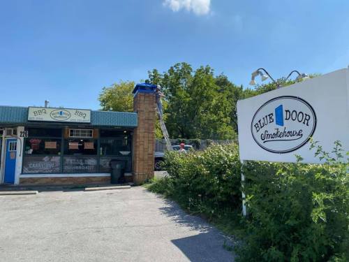Exterior of Blue Door Smokehouse, featuring a sign and a building with a green awning on a sunny day.