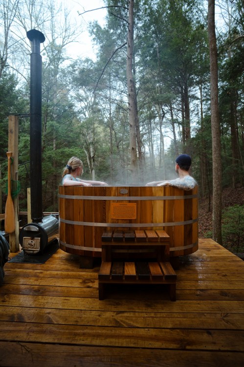 Two people relax in a wooden hot tub surrounded by trees, with steam rising in a serene, forested setting.