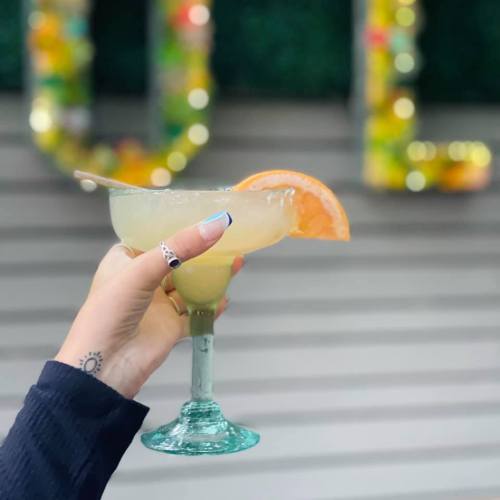 A hand holding a margarita glass with a slice of orange, set against a colorful background.