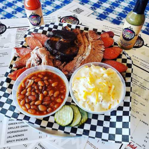 A platter of barbecue featuring smoked meats, baked beans, coleslaw, pickles, and sauces on a checkered tablecloth.