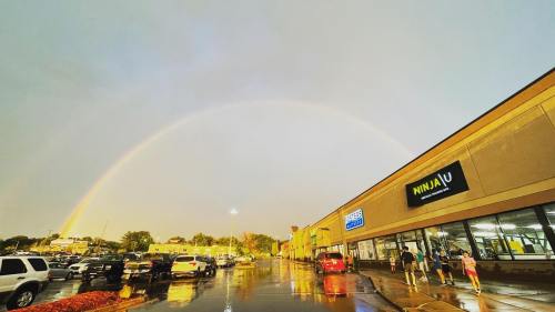 A vibrant rainbow arcs over a shopping plaza after rain, with cars parked and people walking nearby.