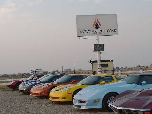 A row of colorful sports cars parked in front of a sign for Sweet Water Smoke, with a cloudy sky in the background.
