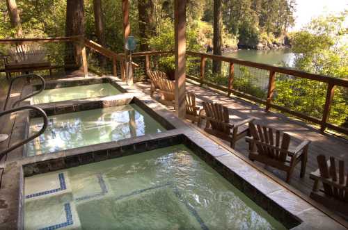 Two hot tubs on a wooden deck overlooking a serene lake, surrounded by trees and outdoor seating.