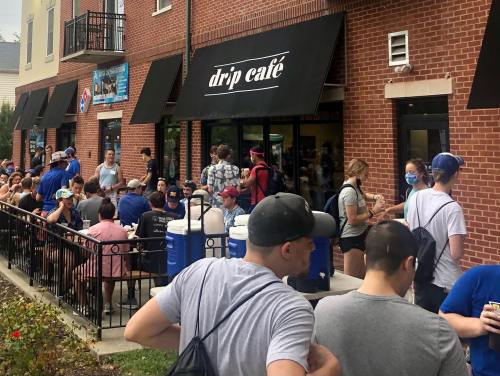 A crowd of people waiting outside Drip Café, with some seated and others in line, on a sunny day.
