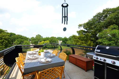A spacious balcony with a dining table, chairs, a grill, and a wind chime, surrounded by greenery and blue skies.