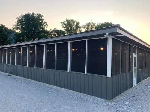 A modern building with a screened porch, surrounded by gravel and trees, under a sunset sky.