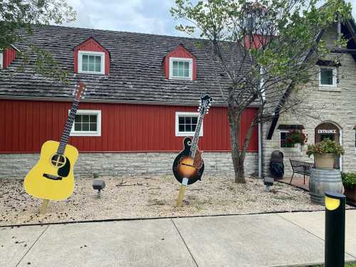 Two large guitar cutouts stand in front of a red and stone building, surrounded by gravel and greenery.