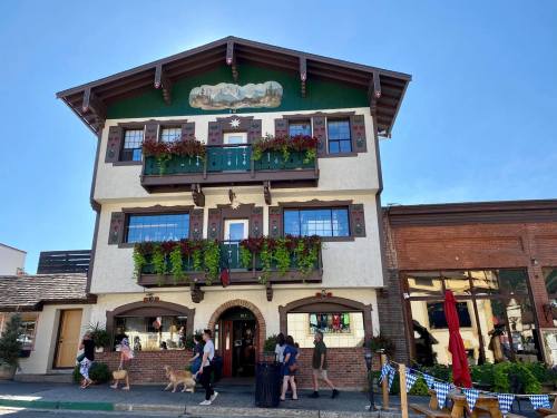 A charming, multi-story building with flower-filled balconies, people walking by, and a clear blue sky above.