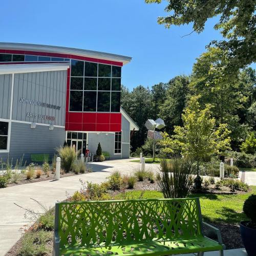 A modern building with large windows, surrounded by greenery and a pathway, under a clear blue sky.