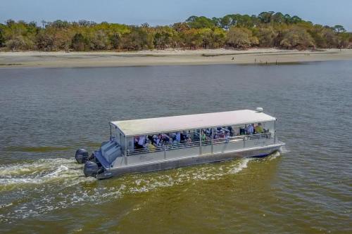 A tour boat with passengers glides through a calm river, surrounded by trees and a sandy shore in the background.