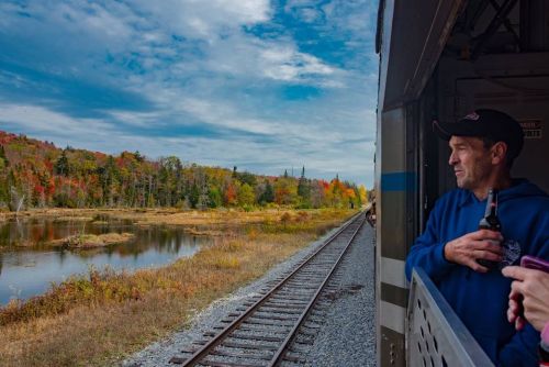 A man leans out of a train window, holding a drink, with colorful autumn trees and a serene lake in the background.