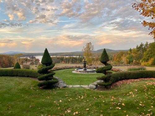 A scenic garden with manicured hedges, a fountain, and a view of rolling hills under a colorful sunset sky.