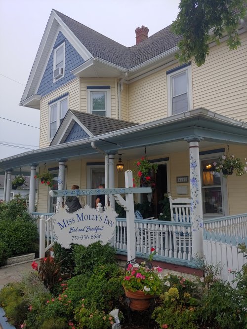 A charming yellow Victorian house with blue accents, featuring a sign for "Miss Molly's Inn" and colorful flower beds.