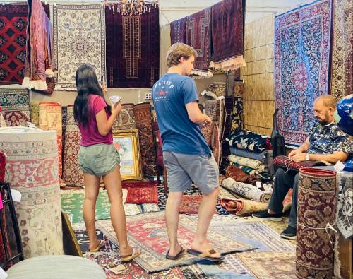 A young woman and man browse rugs in a vibrant market stall, while an older man sits nearby, working on a carpet.