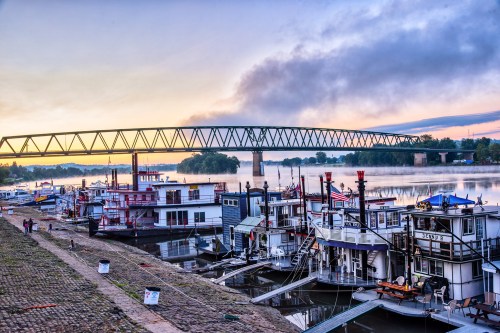 Sunrise over a river with boats docked and a bridge in the background, creating a serene waterfront scene.