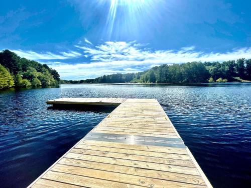 A wooden dock extends into a calm lake, surrounded by lush greenery under a bright blue sky.