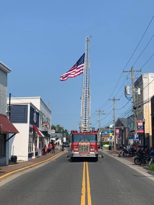 Fire truck with a raised ladder displaying an American flag, set on a street lined with shops and clear blue sky.