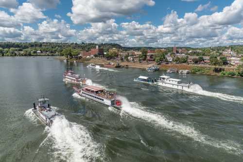 Aerial view of multiple boats navigating a river with a scenic town and cloudy sky in the background.