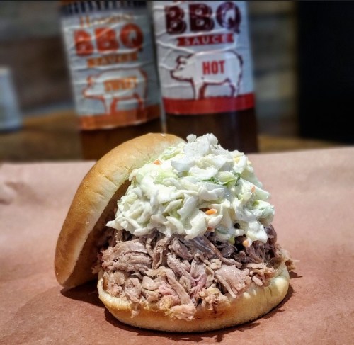 A pulled pork sandwich topped with coleslaw, with bottles of BBQ sauce in the background.