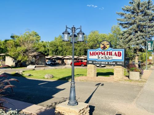 Sign for Moosehead Lodge with a lamp post, surrounded by greenery and cabins in the background. Clear blue sky above.