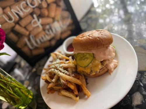A juicy burger with sauce and pickles, served with seasoned fries and a small bowl of ketchup on a white plate.