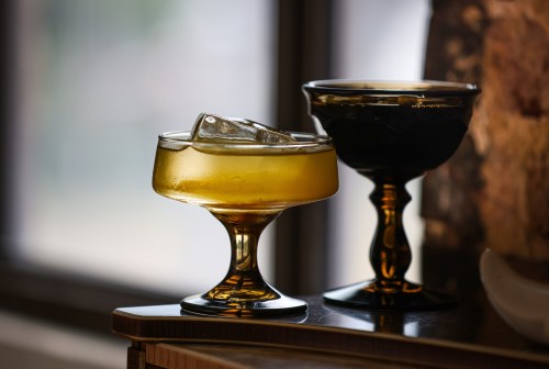 Two elegant glasses on a table, one filled with a golden drink and ice, the other empty and dark. Natural light in the background.