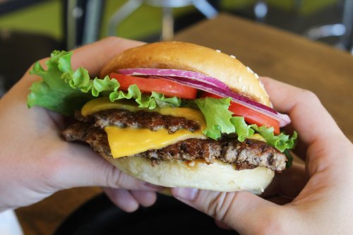 A person holding a cheeseburger with lettuce, tomato, onion, and a sesame seed bun.