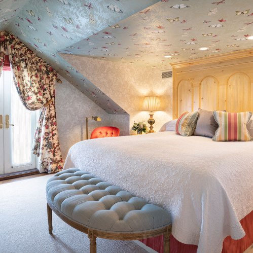 Cozy bedroom with a patterned ceiling, floral curtains, a large bed, and a tufted bench at the foot.