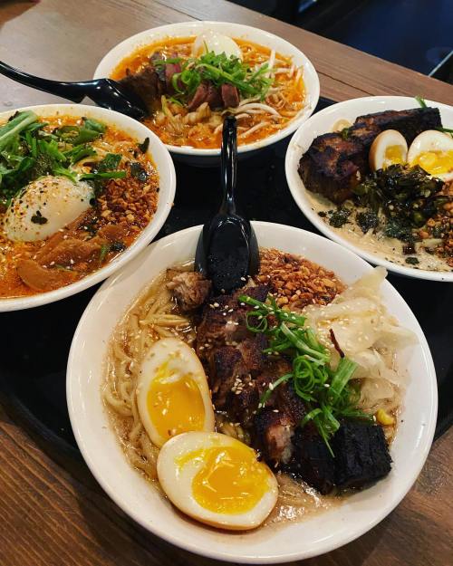 Four bowls of ramen with various toppings, including eggs, pork, and vegetables, arranged on a wooden table.