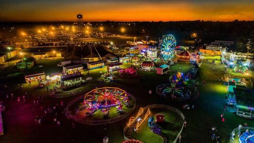 Aerial view of a vibrant carnival at dusk, featuring rides, games, and colorful lights against a sunset backdrop.