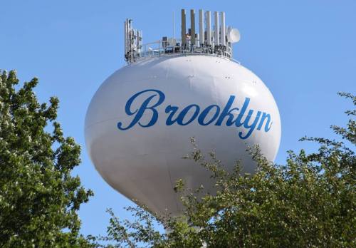 A water tower with the word "Brooklyn" painted in blue, surrounded by green trees against a clear blue sky.
