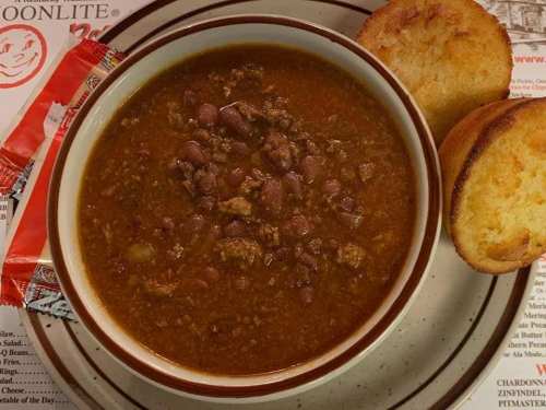 A bowl of chili with ground meat and beans, served with two pieces of cornbread on the side.